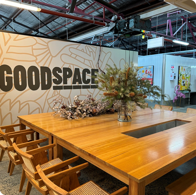 Wooden table with a central planter, surrounded by chairs, against a wall featuring the word "GOODSPACE" in bold letters, highlighting a modern workspace atmosphere.