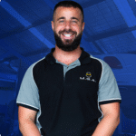 Mechanic smiling in a workshop, wearing a branded shirt from MGA Motors, showcasing expertise in automotive services and customer satisfaction.