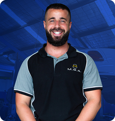 Smiling mechanic wearing a black and gray polo shirt with "M.G.A." logo, standing in a workshop environment, representing client testimonial for Perceptiv Media's SEO services for mechanics.