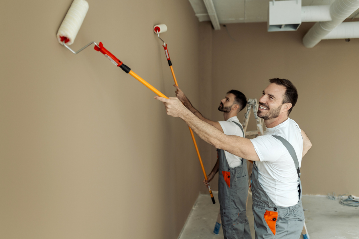 Two painters applying paint to a wall using long rollers, showcasing teamwork and professional painting techniques in a modern interior setting.